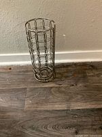 Front and side views of a silver metal wire toilet paper holder placed on a wooden floor against a wall.