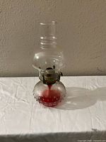 Oil lamp showing full view with red hobnail glass base, metal burner, and clear glass chimney on white cloth background