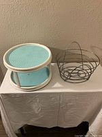 Metal wire basket next to two-tier oval Lazy Susan with white frame and blue platforms on white cloth-covered surface against beige wall