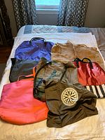 Eight assorted tote bags laid out on a bed showing different colors and materials, including canvas, polyester, and faux leather with various straps and pockets.