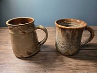 Two handmade ceramic mugs on a wooden surface, showing front and side views highlighting shape and glaze details.