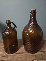 Photo showing two vintage glass bottles: a brown Javex bottle with handle on left, and a tall tall brown swirly patterned glass bottle with cork on right. Bottles placed on wooden surface against neutral wall.