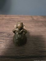Front view of small brass bell shaped like a lady with bonnet hat and hands on hips on wooden surface.