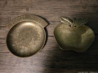 Top view of two brass coin trays: round tray embossed with "Pocket Change" and apple-shaped tray with leaf decoration, showing surface wear and tarnish.