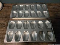 Two vintage metal baking pans, each with 12 classic madeleine shell-shaped molds, placed on a wooden surface for display.