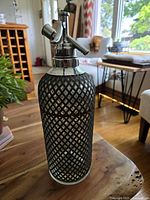 Front angle showing full vintage seltzer bottle with diamond patterned cover and metal siphon top.