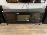 Front view of black wooden TV stand with built-in electric fireplace and glass panel side cabinets. Remote control placed on top.