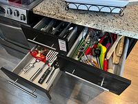 Three kitchen drawers opened showing the contents of cutlery and kitchen utensils including knives, whisks, peelers, and other tools.
