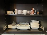 Cupboard shelf with white ceramic bowls, mugs with various designs, glass measuring cups, and glass dishes.