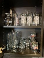 Cabinet shelf with variety of stemmed wine glasses, tall and short clear drinking glasses, and decorative holiday-themed glasses.