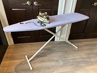 Iron and ironing board placed upright on wood floor in front of dark double doors.