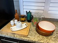 Wide view of the kitchen items on a countertop including teapot, cutting board, bowl, pitchers and other pottery items