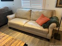 Front view of beige leather three seat couch with two seat cushions, three back cushions, one orange throw pillow and dark green throw blanket on right armrest.