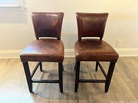 Two brown leather bar stools or chairs with padded seats and backs, set on wooden legs, shown from the front, against a white wall and light wood floor.