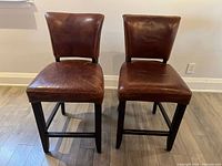Front view of two brown leather stools with wooden legs and cushioned seats and backs.