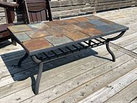 Rectangular patio coffee table with multi-colored stone tile top showing natural earthy tones and black metal legs on a wooden deck.