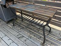 Front angle view showing rectangular stone tile tabletop in patterned layout, black curved metal legs and lower metal slatted shelf on wooden deck.