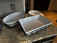 Two white ceramic serving dishes, one rectangular and one oval, each in black metal holders with curved decorative designs, placed on granite countertop.