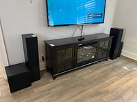 Three audio components against wall under mounted TV: left stack shows subwoofer below floorstanding speaker, right shows matching floorstanding speaker. Remote on credenza.