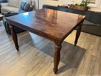 Square wooden table in a room, showing top and legs with dark wood and polished finish.