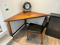 Photo showing the wooden corner triangle table with a black-framed round table clock on top and a black leather chair next to it.