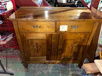 Front view of medium brown wooden sideboard showing two drawers at top and double cabinet doors below with carved details. Sideboard has curved legs and traditional style.