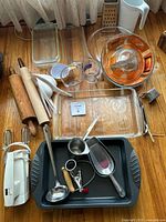 Lot of assorted baking and kitchen tools laid out on wooden floor under window with white sheer curtains. Multiple glass dishes, measuring cups, hand mixer and utensils visible.