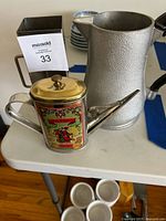 Photo showing assorted metal pitchers and jugs including a textured silver-tone pitcher, a small teapot-style jug with brass lid and vintage label, and a square metal pitcher with handle.