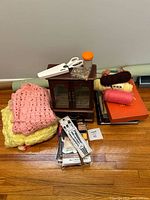Photo showing the full lot with pink and yellow crocheted afghans, wooden cabinet, orange pin container, unused scissors, yarn spools, books, packaged sewing needles, and zippers.