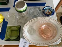 Nine-piece lot displayed on a table showing assorted mid-20th century glassware and ceramics including depression glass items and decorative ceramic plates and goblet.