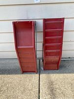 Pair of two red metal car ramps positioned upright on pavement against siding wall, showing front surface with horizontal ridges for tire grip.