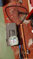 Photo showing three fishing rods, a fishing net, a large tackle box, a smaller container with hooks, and a brown carrying tube on floor.