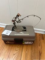 Top view showing brown Flambeau fishing tackle box with black latch, fishing-themed metal sculpture placed on top with fisherman holding fishing rod.