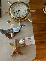 Brass nautical ship wheel clock mounted on wall with white dial and 24-hour numbering shown with surrounding décor including metal bird sculpture, starfish and sea shell.