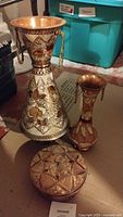 Photo showing two hand-engraved brass vases and a lidded box placed on a surface with background storage bins.