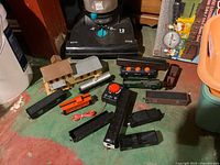 Photo showing multiple HO scale train cars including orange locomotive, black freight cars, two small buildings, and a black Mehano transformer/controller with red dial on a green floor.