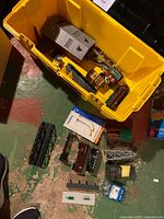 Yellow plastic bin containing mixed model train buildings and accessories including gray building with gabled roof, multiple small buildings, and a black bridge section on floor outside bin.