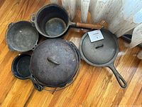 Photo showing 10 cast iron cookware pieces including several skillets, pots, and two Dutch ovens with lids, one pot with wooden handle visible.