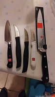 Top view of five knives arranged side by side on a patterned surface including two Zwilling paring knives, one R & R International paring knife, one Wusthof chef's knife out of packaging, and one Wusthof chef's knife in packaging.