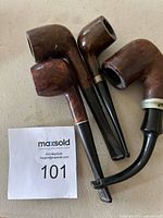 Four wooden and plastic smoking pipes arranged on a flat surface with a MaxSold label number 101. Pipes vary in shape and show metal accent rings on some parts.
