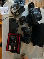 Top view of vintage cameras and binoculars on white table showing Ricoh camera with case, Argus camera, binoculars and carrying cases.