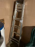 Full view of aluminum ladder leaning against wall near water heater and green chair; showing six step rungs and black plastic top tray.