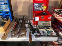 Photo of a table displaying mixed hand tools including saws, a level, hatchet, mallet, pipe wrench, hacksaw, and a red metal toolbox with additional accessories inside