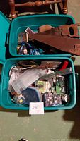 Two large blue plastic storage bins filled with assorted hand and power tools, hardware kits, fishing line, and various small accessories. Top bin contains rusty hand saws and other hand tools, bottom bin contains hardware kits, small packaged parts, and plastic organizers.