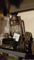 Photo of two vintage metal lanterns, one clear glass lantern chimney, electric iron with cord, and wooden plane with knob handle on shelf.