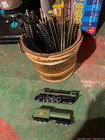 Photo showing green locomotive and tender beside a wooden bucket filled with black model train track pieces.