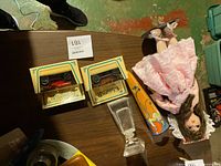 Porcelain doll lying on table with two boxed die-cast cars and a glass decanter in a triangular arrangement.