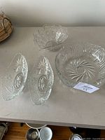Four clear pressed glass serving bowls and dishes on a beige surface, including two oval dishes, one large round bowl, and one smaller scalloped bowl with starburst and geometric pressed glass designs.