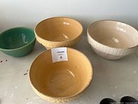 Four ceramic mixing bowls on a white surface with a MacSold lot label visible. Includes two yellow textured bowls, one cream bowl with exterior pattern, and one green bowl.