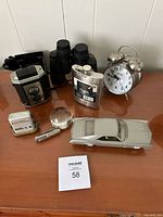 Photo showing Tasco Sonoma binoculars, Kodak Brownie Reflex camera, metal car model, alarm clock, flask, magnifying glass, and calendar on a wooden surface.